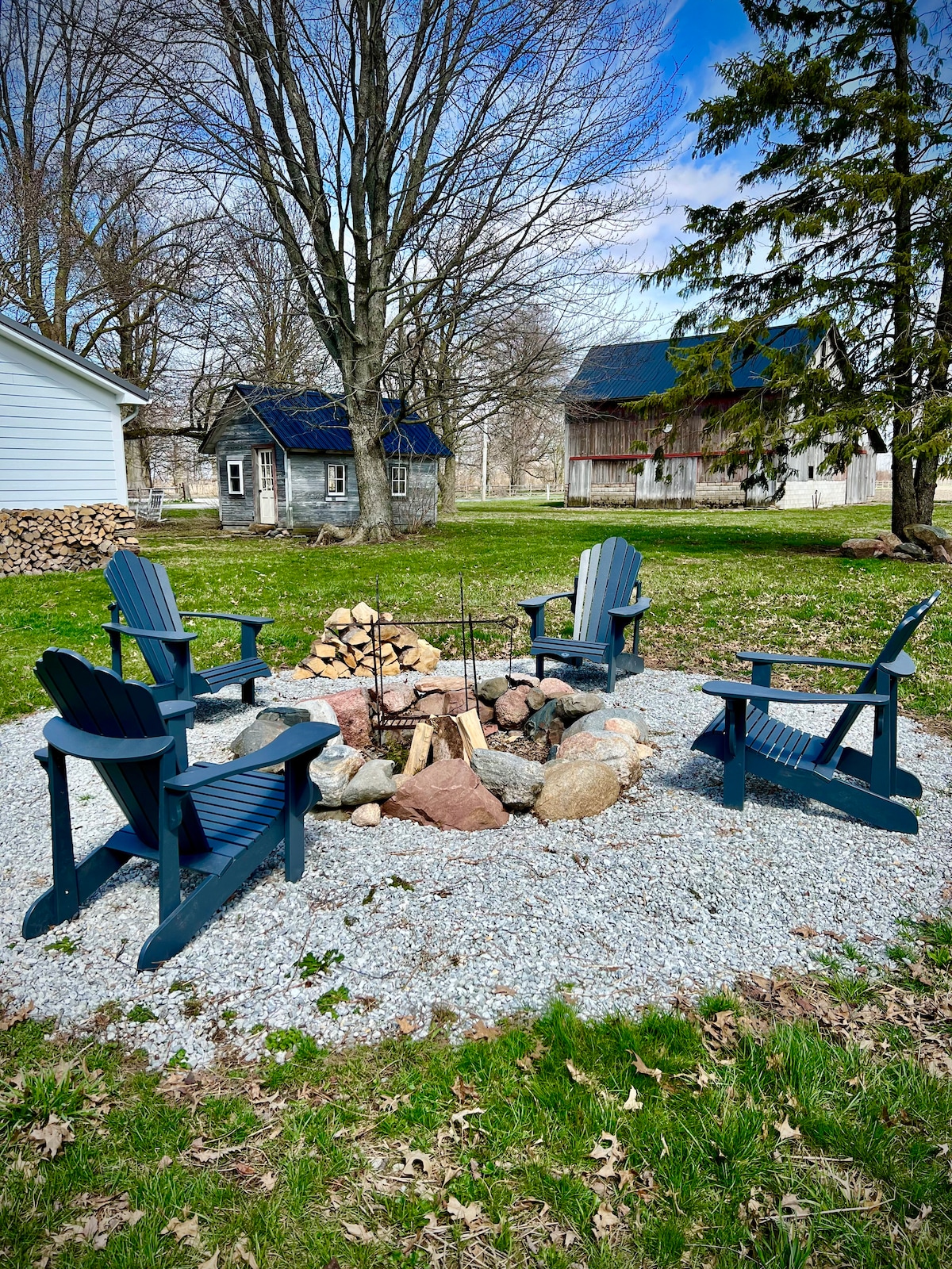A fire pit area is surrounded by four wooden adirondack chairs on a gravel base. Nearby, stacks of firewood are neatly organized, and rustic barn structures are visible in the background. Tall trees provide natural shade in the open space.