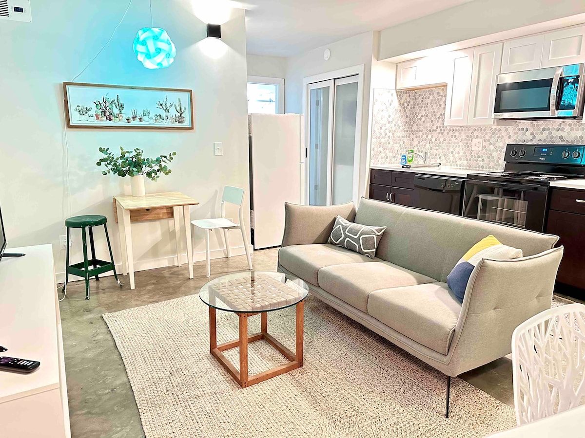 A cozy living area features a light gray sofa accompanied by a round glass coffee table. A small dining table with two chairs sits nearby. The kitchen, visible in the background, includes dark cabinetry and modern appliances, while decorative plants add a touch of greenery.