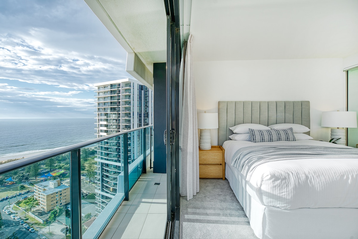A serene bedroom area features a spacious bed adorned with soft linens and decorative pillows. Floor-to-ceiling glass doors open to a balcony, offering expansive views of the ocean and the surrounding coastline, while natural light brightens the room.