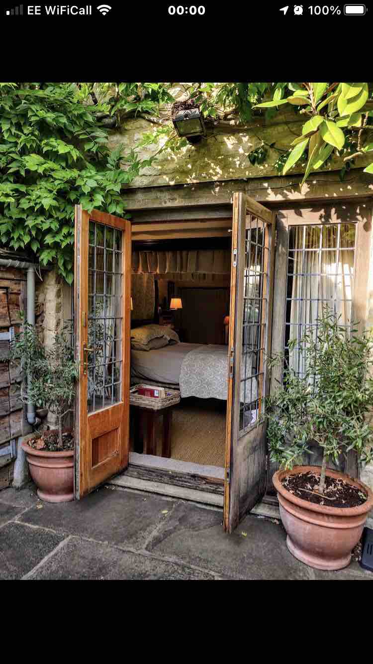 Double French doors framed by lush greenery open to reveal a cozy bedroom. The inviting space features a neatly made bed, soft lighting, and warm tones, with terracotta pots on either side adding a touch of nature.