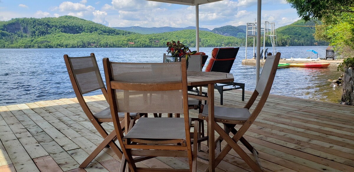 A wooden deck overlooks the lake, featuring a round dining table surrounded by four chairs. Nearby, a lounge area offers two chairs with colorful cushions. The scene is framed by tranquil waters and lush green mountains in the background under a partly cloudy sky.