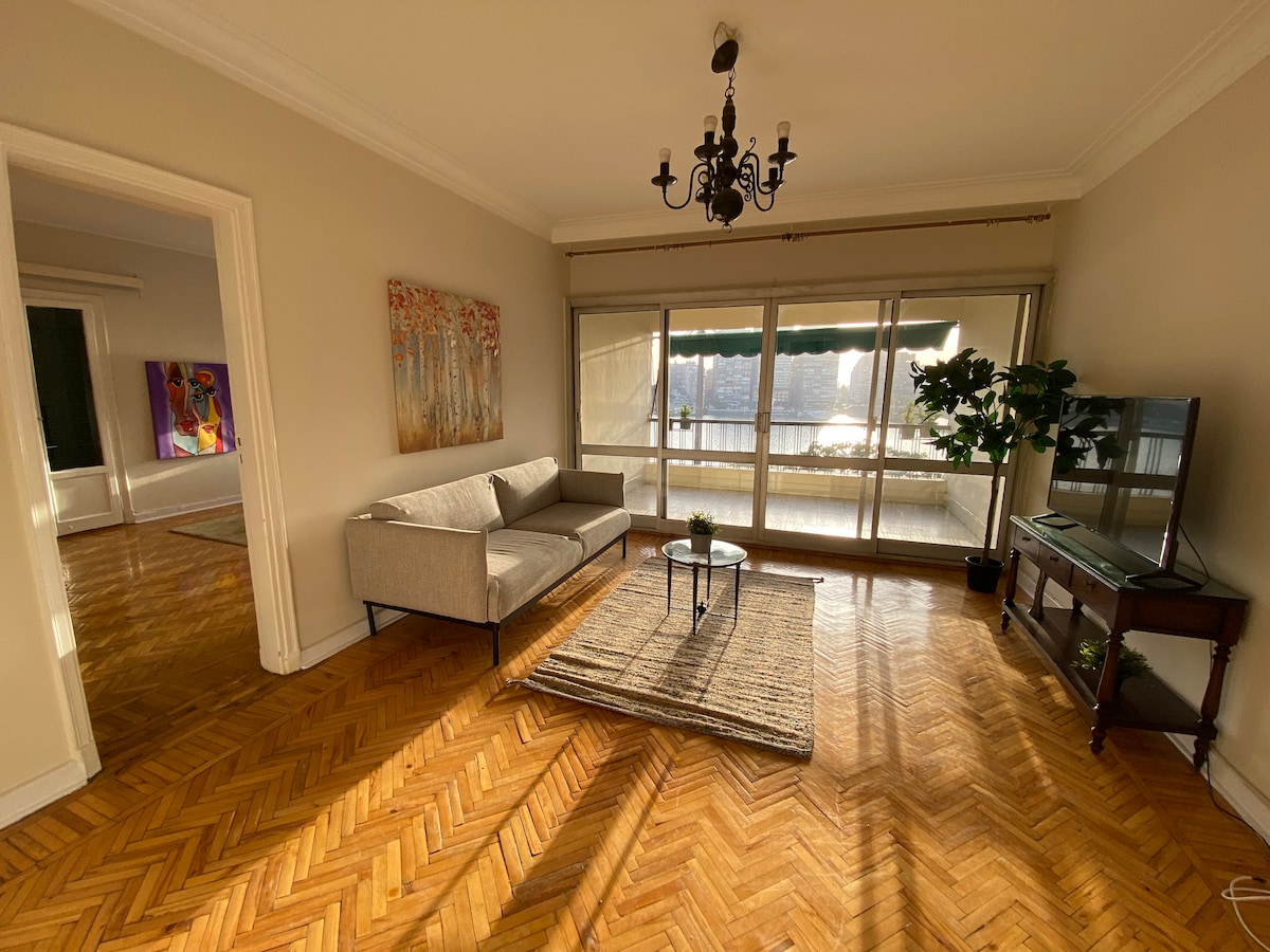 A bright living area features a two-seated couch and a television on a console. Floor-to-ceiling windows allow natural light to fill the space, revealing the scenic views outside. A decorative plant adds a touch of greenery, and a woven rug sits on the polished hardwood floor.
