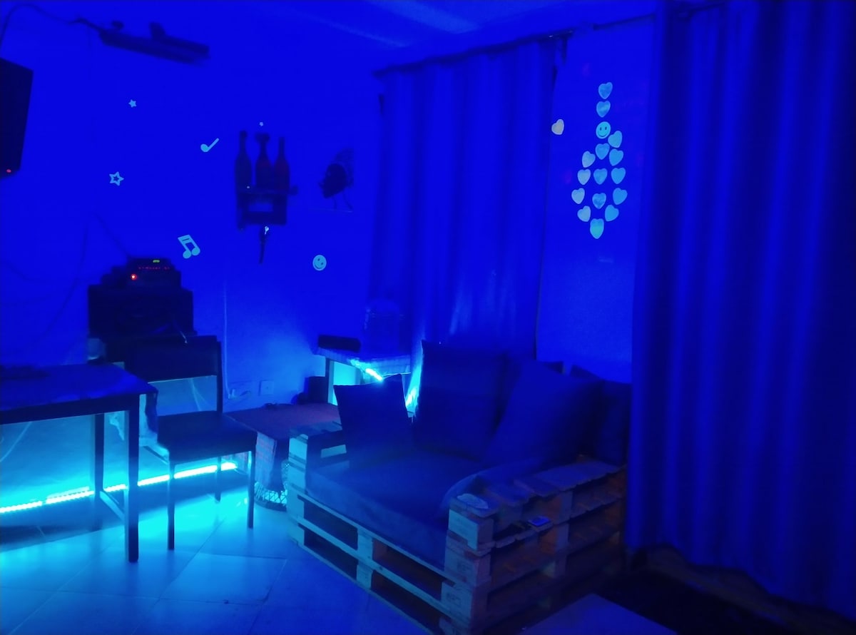 A serene space is illuminated in blue light, creating a calm atmosphere. A couch made of pallets is positioned against a wall, complemented by a table and decorative elements on the walls. Soft curtains frame the room, adding to the tranquil environment.