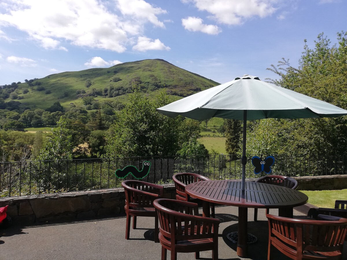 A spacious outdoor seating area features a round wooden table surrounded by several chairs. A large umbrella provides shade while verdant hills rise majestically in the background, under a blue sky adorned with fluffy clouds. Lush greenery frames the setting.