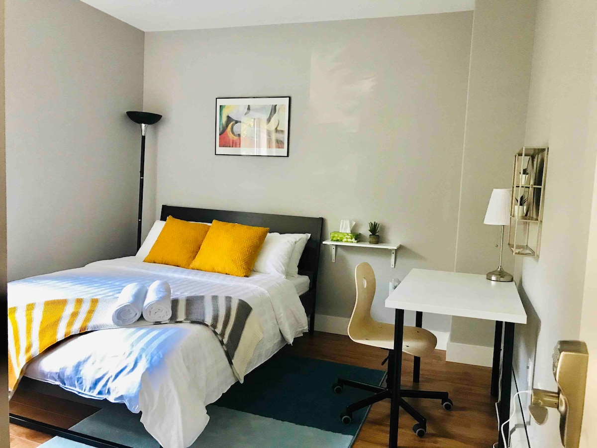 A well-lit bedroom displays a spacious bed with crisp white linens and bright yellow accent pillows. A small desk and chair are positioned near a window, while a stylish floor lamp adds additional light. A grey area rug introduces a soft touch to the wooden floor.