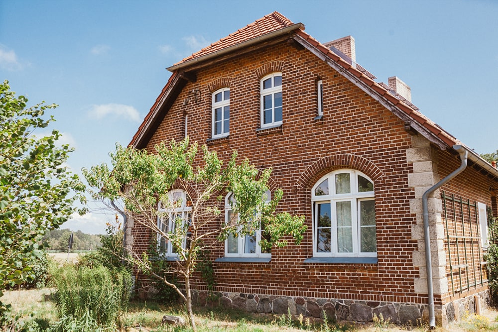 The exterior of the renovated old schoolhouse is showcased, featuring traditional red brick walls and characteristic arched windows. Lush greenery is positioned around the building, with a tree adding natural elements to the serene setting. The surrounding landscape includes open fields and distant trees.