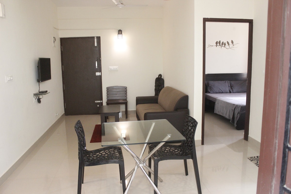 A living area is visible, featuring a brown sofa and two black woven chairs around a glass table. A television is mounted on the wall, while a doorway leads to a bedroom in the background. Soft lighting provides a calm ambiance.