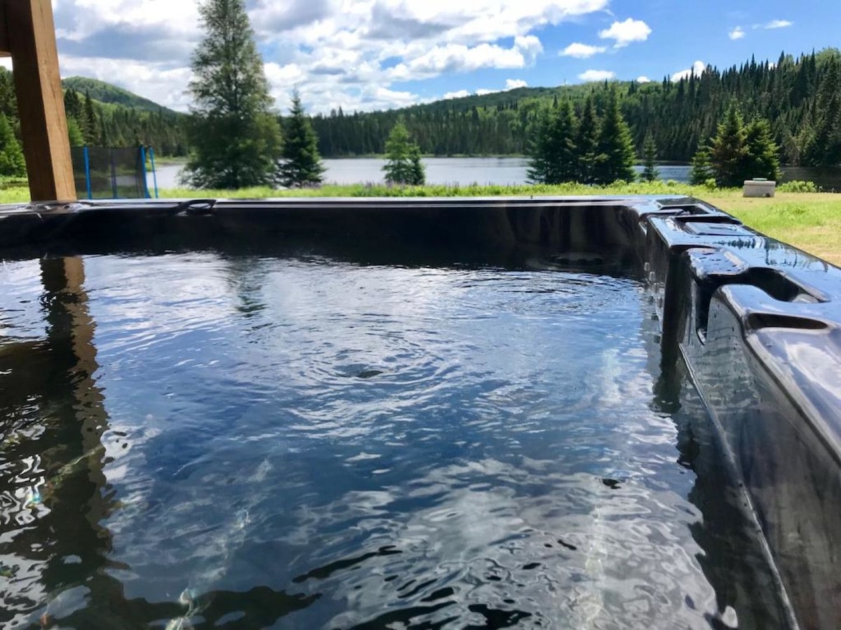 A hot tub is positioned on the deck, featuring clear water that reflects the sky. In the background, a serene lake is surrounded by lush green trees and rolling hills, providing a tranquil escape.