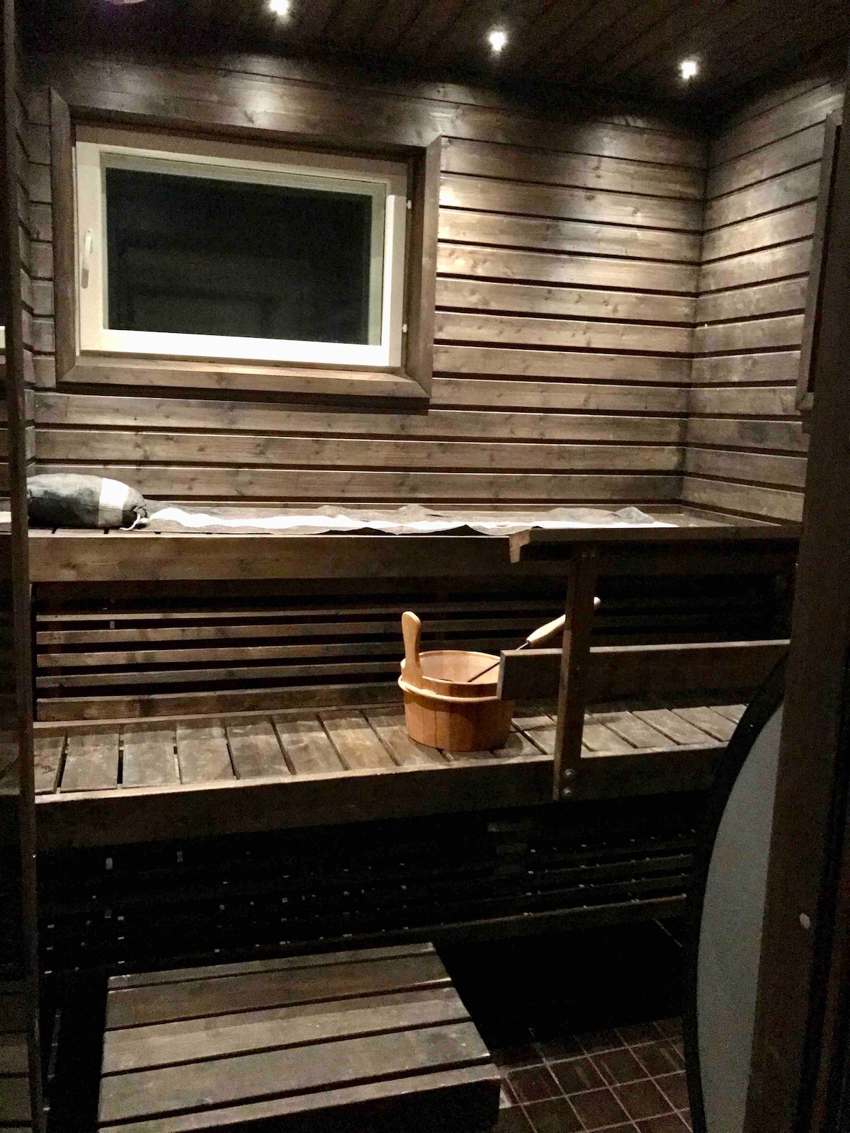 A wooden sauna interior features a series of horizontal benches made of slatted wood, creating a warm and natural atmosphere. A small wooden bucket sits on the lower bench, and a window allows soft light to enter the space, enhancing the cozy environment.