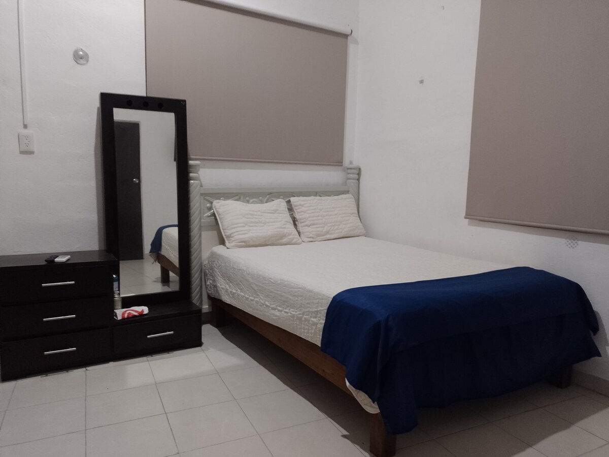 A sleeping area features a spacious bed with a blue bedspread positioned against a light-colored wall. A full-length mirror stands next to a dark wooden dresser with several drawers. Natural light enters from a nearby window, partially covered by beige shades.