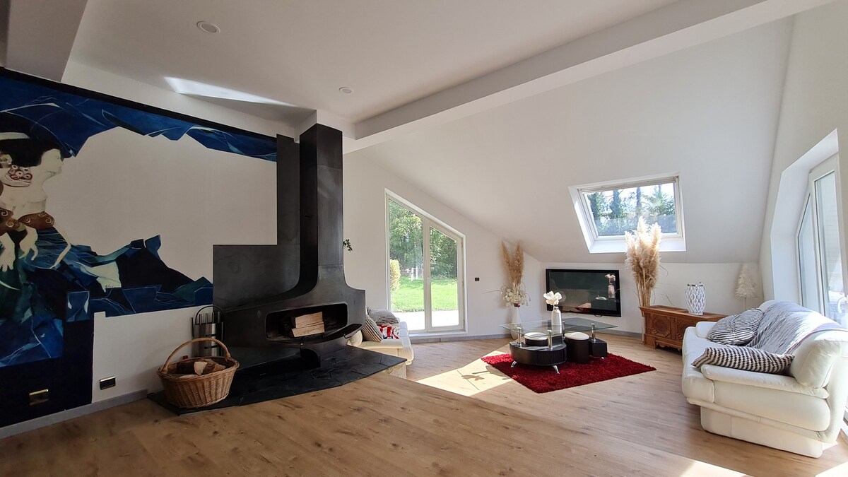 A spacious living area features a modern fireplace on a dark wall, complemented by large windows that bring in natural light. A comfortable white sofa is arranged with a round table, while a vibrant red rug adds warmth to the wooden flooring.