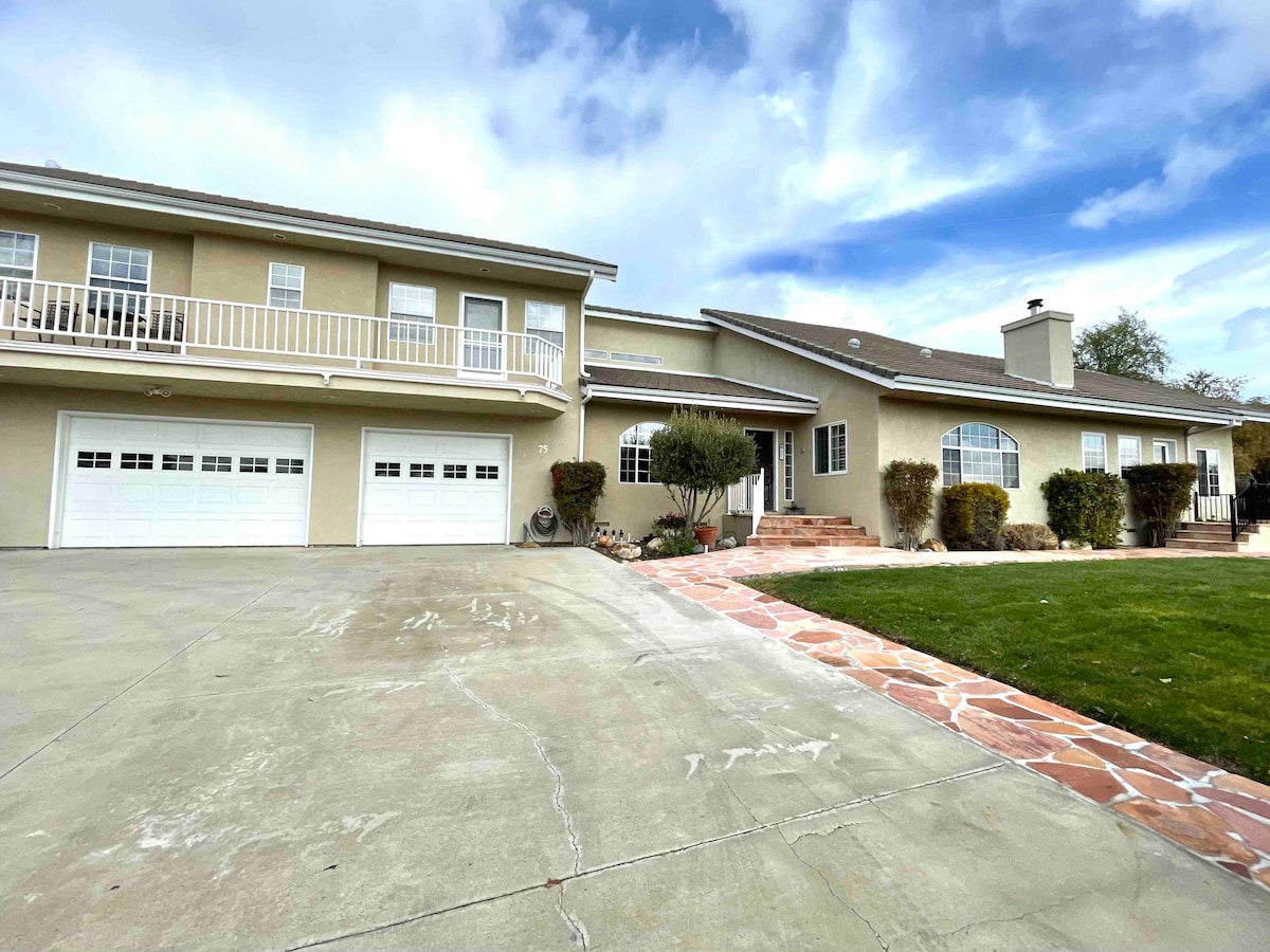 A two-story home is presented with a light exterior, featuring a spacious front driveway and a large, grassy area. The entrance is framed by neatly trimmed bushes, while a balcony above overlooks the front. Multiple garage doors are visible, enhancing accessibility.