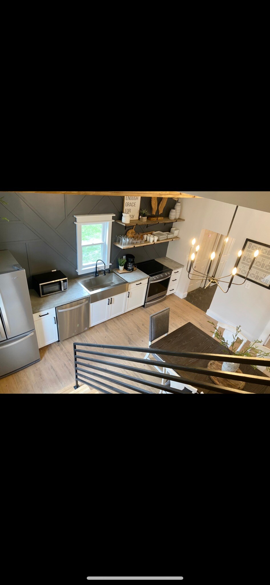 A modern kitchen with stainless steel appliances is visible, featuring a microwave, coffee maker, and sleek cabinetry. Natural light enters through a window, illuminating the countertop. A dining area with a dark wooden table is positioned underneath a stylish chandelier, completing the inviting space.