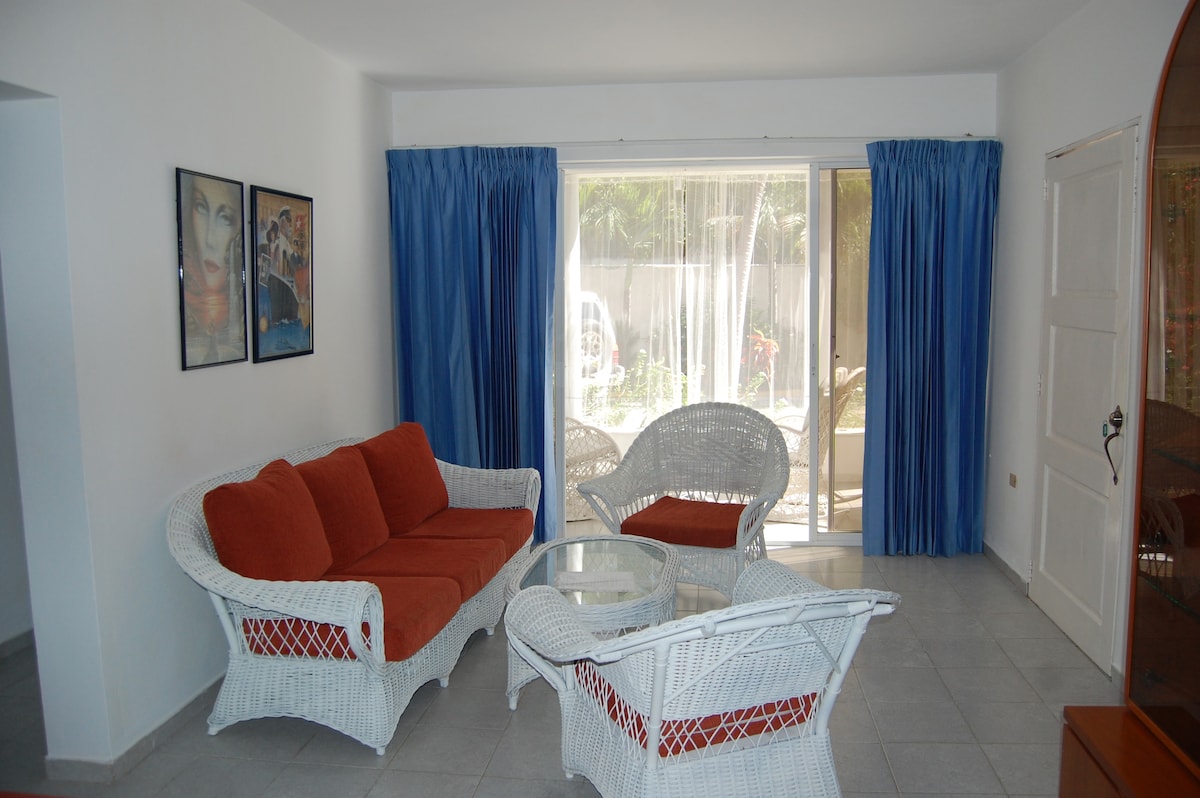 A light-filled living area features white wicker furniture, including a sofa with orange cushions and two armchairs. A glass table sits in the center. Large glass doors offer views into a garden space, while blue curtains add a touch of color to the white walls.