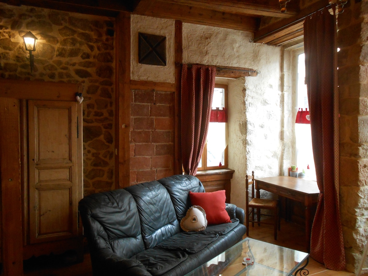 A rustic living area features a leather sofa next to wooden furniture, with a small table by the window. Natural light filters through the window, adorned with red curtains, while stone and wooden walls add character to the space.