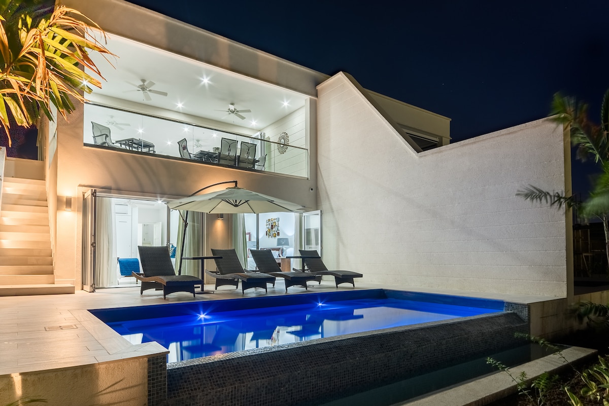The exterior features a private pool with a smooth blue surface, accompanied by comfortable loungers. The modern structure showcases large glass walls that offer a view of the open-plan interior space, illuminated softly against the night sky.