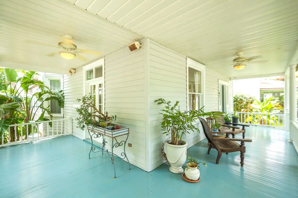 A wrap-around porch features a light blue floor and white wooden walls, providing a refreshing space to relax. Potted plants are arranged on either side, and two wooden chairs offer comfortable seating. Ceiling fans are located above, enhancing the tranquil atmosphere.