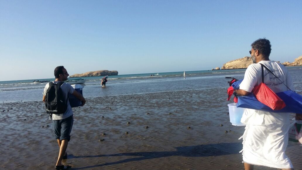 Two individuals walk along a wide, sandy beach, wearing casual attire and carrying buckets and bags. The shallow water glistens under the sun, with a few boats visible in the distance. A rocky formation rises on the shore, set against a clear blue sky.