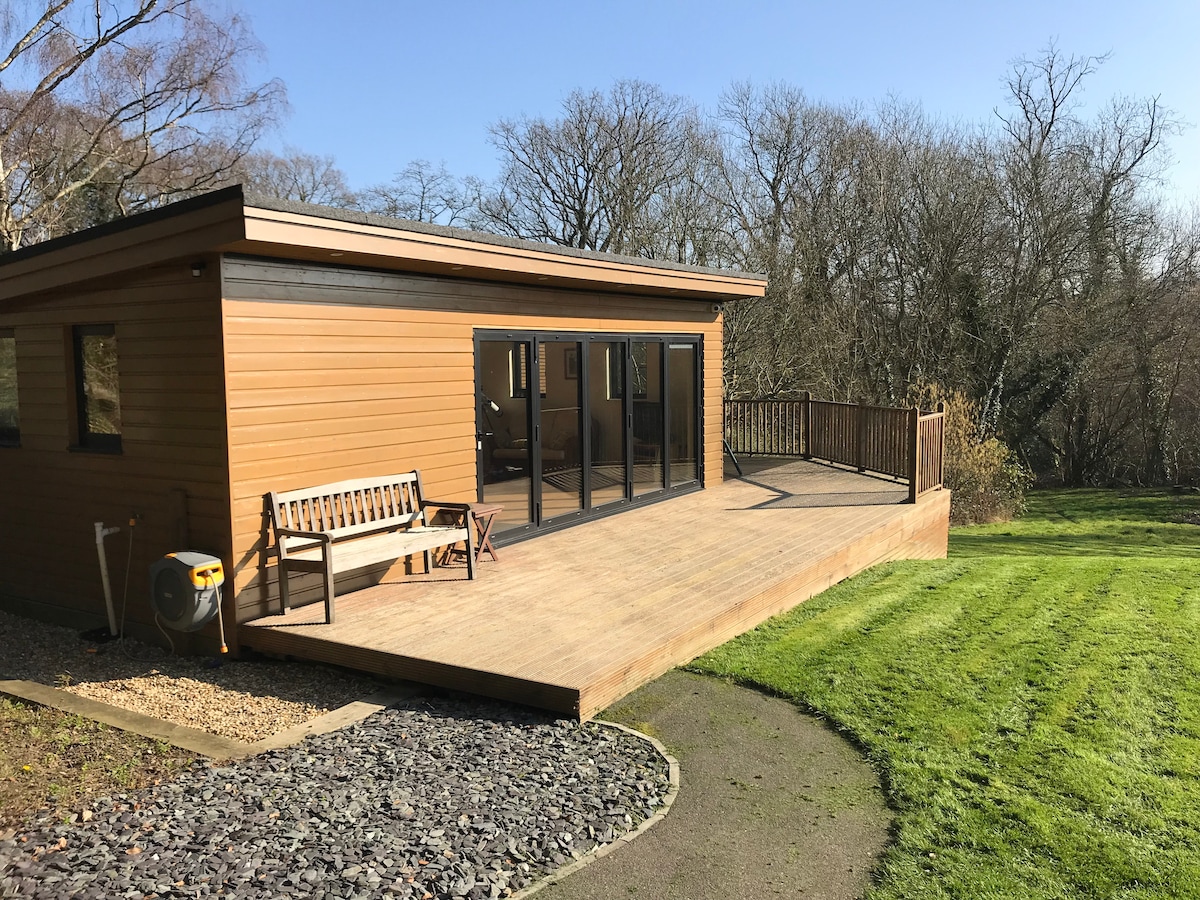 The exterior of a modern Summer House is shown, featuring large glass doors that open to a spacious deck. A wooden bench is positioned on the deck, surrounded by manicured grass and trees in the background, providing a tranquil view of the natural surroundings.