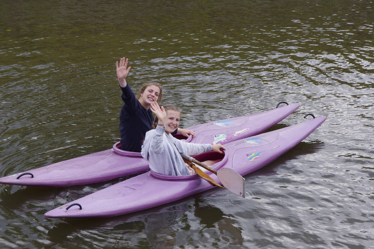 Two individuals are seated in separate pink kayaks on calm water, each raising a hand in greeting. The surrounding water reflects the soft ripples created by their movements, creating a serene atmosphere.