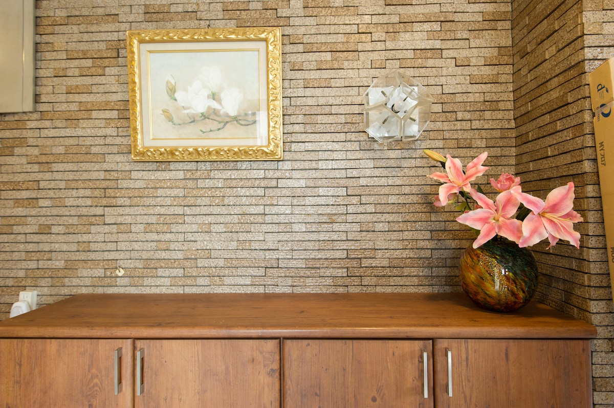 A wooden sideboard is placed against a textured stone wall, complementing the warm tones of the decor. Above the sideboard, a floral painting in a gold frame adds a touch of artistry. A vase filled with pink lilies provides a subtle pop of color.
