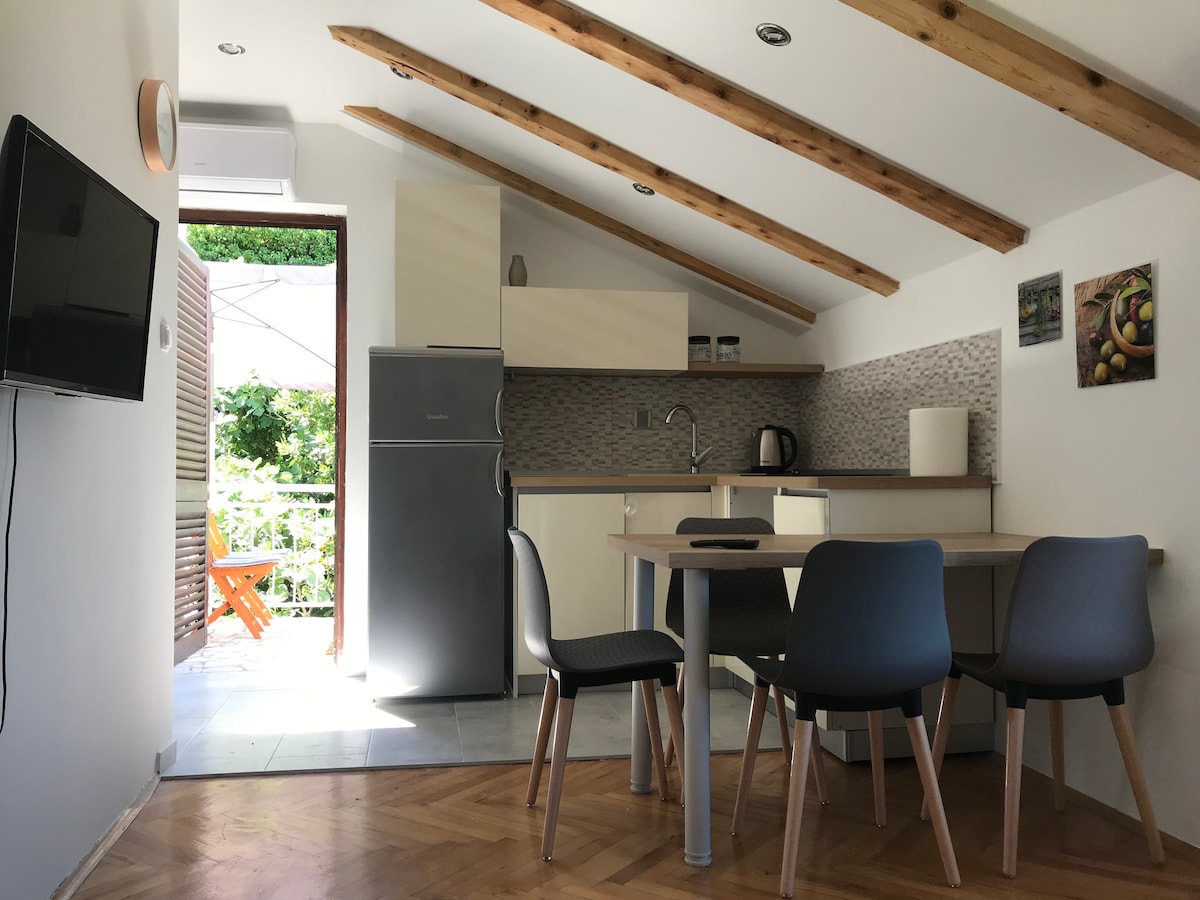 A bright kitchen area is furnished with modern cabinetry and a refrigerator, while a dining table surrounded by chairs occupies the center. Large windows allow natural light to fill the space, creating an inviting environment. An air conditioning unit is mounted on the wall.