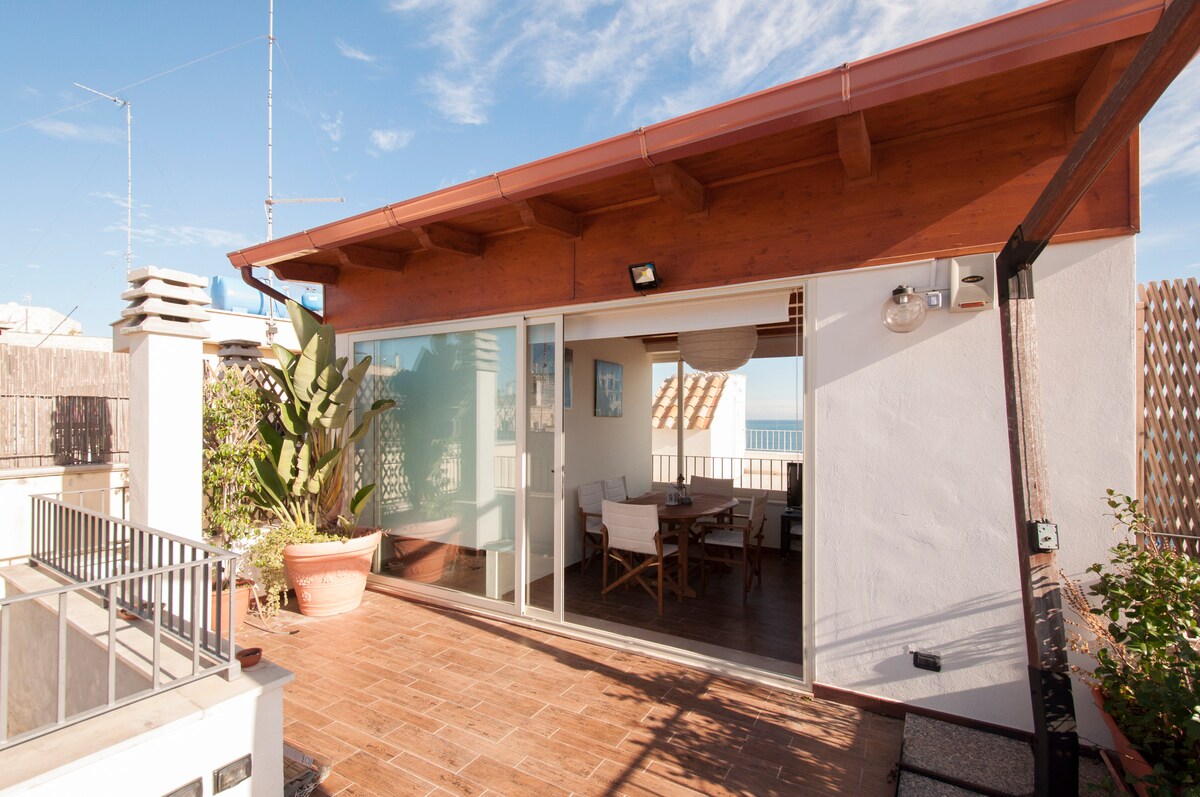 A cozy terrace is adorned with potted plants and a wooden roof, providing an inviting outdoor space. Large glass doors connect the terrace to the interior, allowing for plenty of natural light. Sea views are visible in the distance against a clear sky.