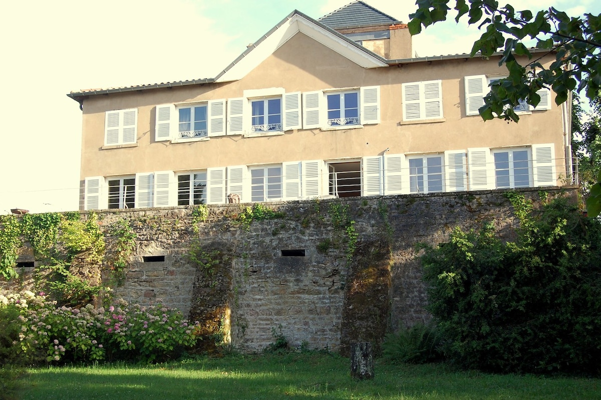 The elegant house features a light-colored facade adorned with numerous white shutters. The structure is set slightly above ground, surrounded by lush greenery and colorful flowering plants, adding a natural charm to the setting. A peaceful atmosphere is suggested by the inviting, well-maintained exterior.