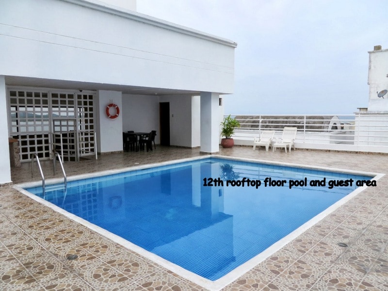 The rooftop pool area is shown with a clear blue pool bordered by a tiled deck. A set of dark chairs and a table are positioned nearby against a white wall, and ocean views are visible in the background under a cloudy sky.