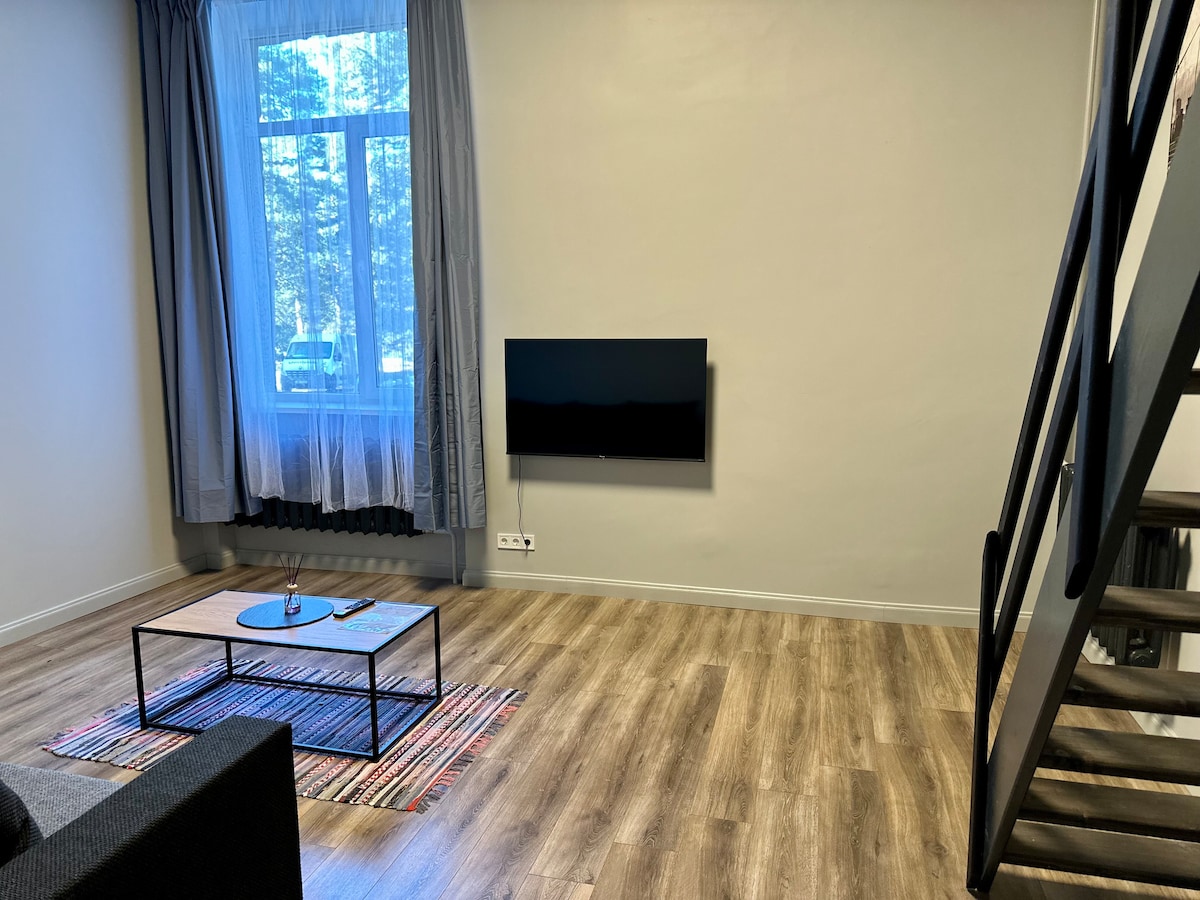 A modern living area features a comfortable sofa and a sleek coffee table placed on a patterned rug. A wall-mounted television is positioned directly across from the seating area. Natural light filters through a large window adorned with sheer curtains.
