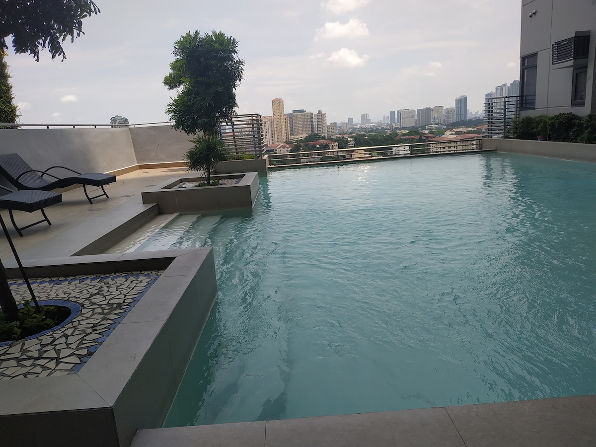 An infinity pool is seen overlooking a city skyline, reflecting the clear sky. Poolside loungers are positioned nearby, surrounded by greenery. The smooth, tiled surface of the pool edges invite relaxation, with modern architectural details enhancing the outdoor space.