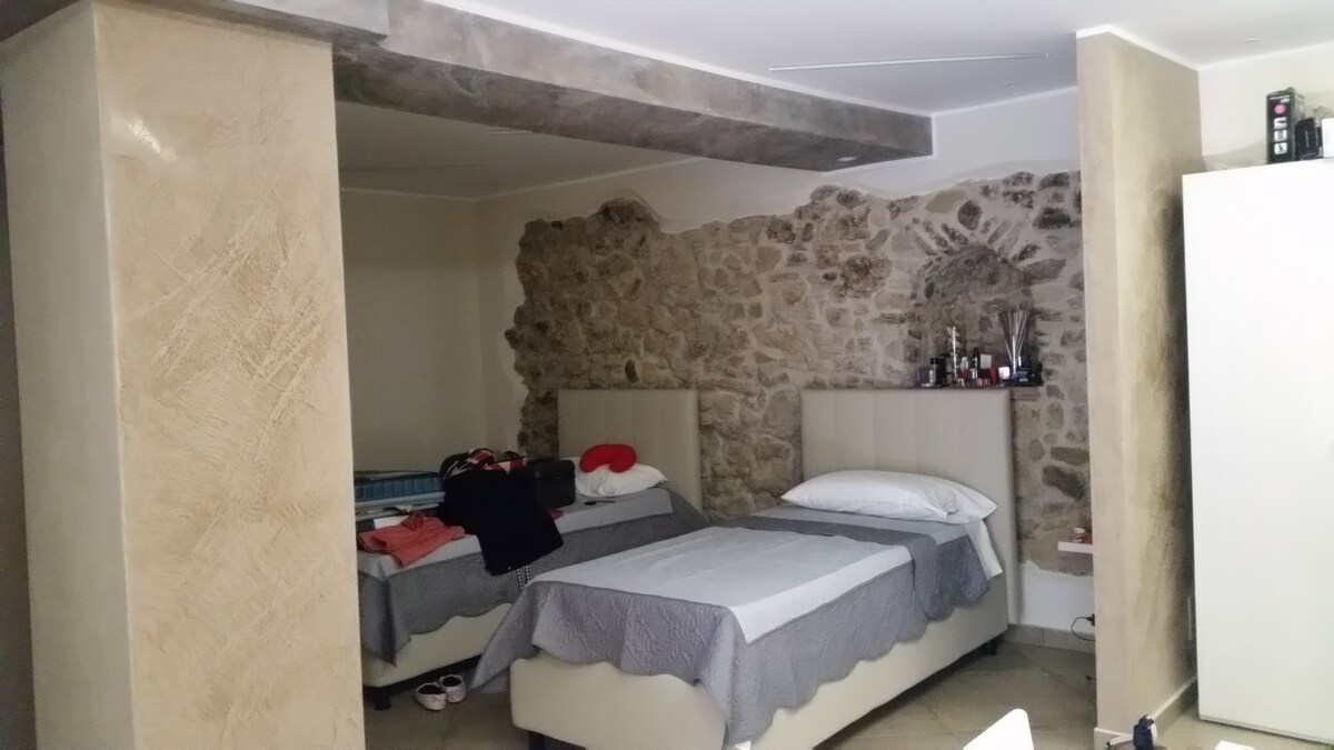 A well-appointed sleeping area features two spacious beds with neutral bedding and complementary headboards. A stone accent wall adds texture, while a minimalistic wardrobe stands nearby. Personal items are visible on the beds, and the warm-toned floor provides a cozy foundation.