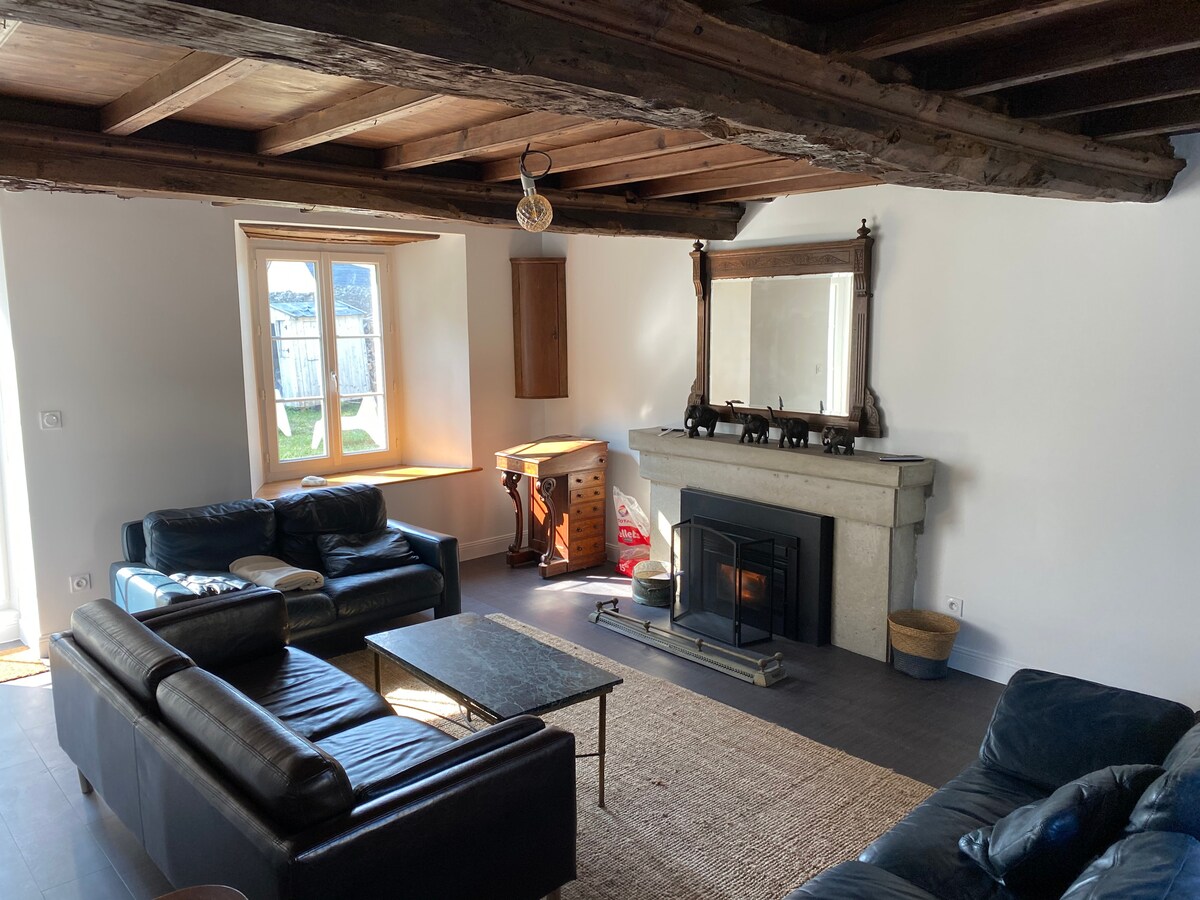 A cozy living room is highlighted by a stone fireplace, providing a focal point against the neutral walls. Comfortable seating is arranged around a coffee table. Natural light filters through the window, illuminating the space, while wooden beams add rustic charm to the ceiling.