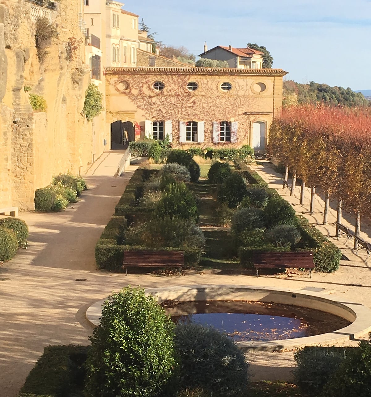 A well-maintained garden is visible, featuring neatly trimmed hedges and a circular water feature. Rows of greenery create a structured layout, while benches provide seating options. The backdrop showcases a charming building with large windows, framed by climbing vines and bordered by autumn-hued trees.