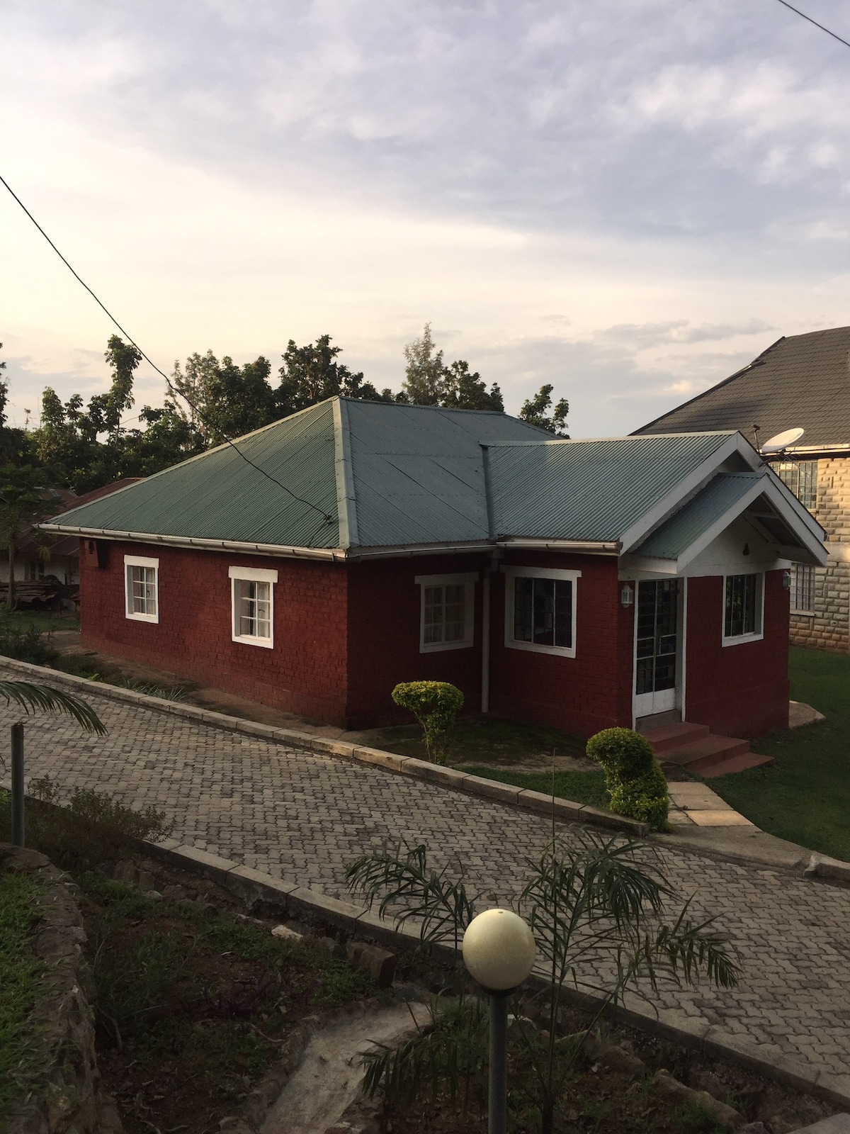 A vintage red house with a sloped metal roof is surrounded by greenery. Decorative shrubs are positioned near the entrance, and the stone-paved path leads to the front door, framed by large windows. The setting is complemented by a peaceful neighborhood backdrop.