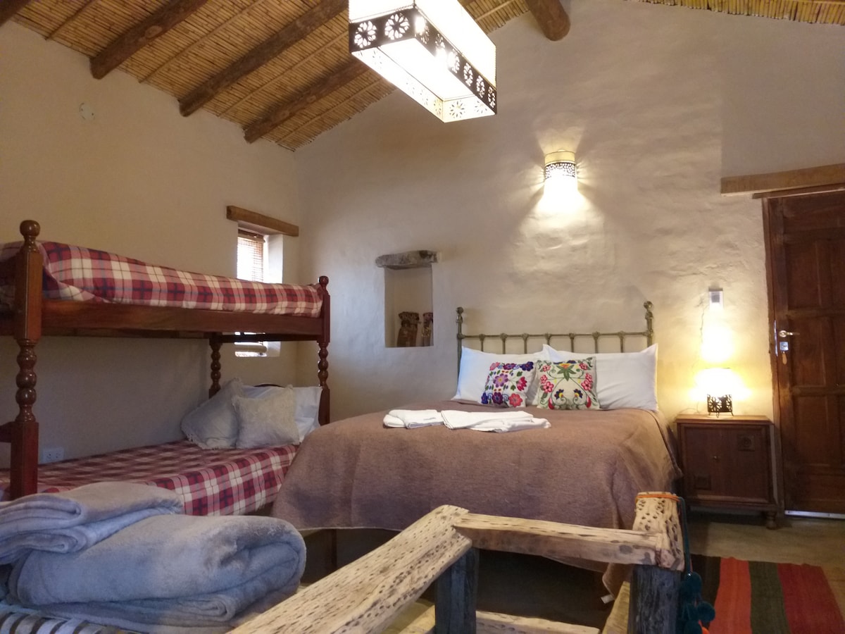 A rustic sleeping area features a spacious bed and a bunk bed, both adorned with simple bedding. Natural textures are highlighted by wooden beams and a lantern-style light fixture. A cozy wooden table sits in the foreground, complemented by a soft rug.