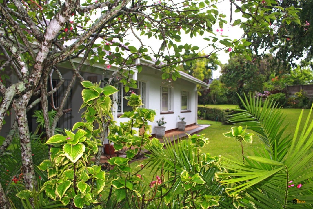 A side view of the house is framed by lush greenery and blooming plants, highlighting the exterior's white walls and wooden elements. The surrounding garden features well-kept grass and ornamental shrubs, creating a serene outdoor environment.