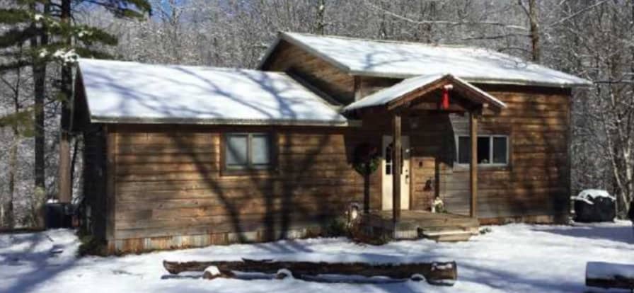 The cabin is set amidst a snowy landscape, featuring a warm wooden exterior and a covered front porch. Shadows of surrounding trees play across the snow-covered ground, enhancing the serene, wintry atmosphere.