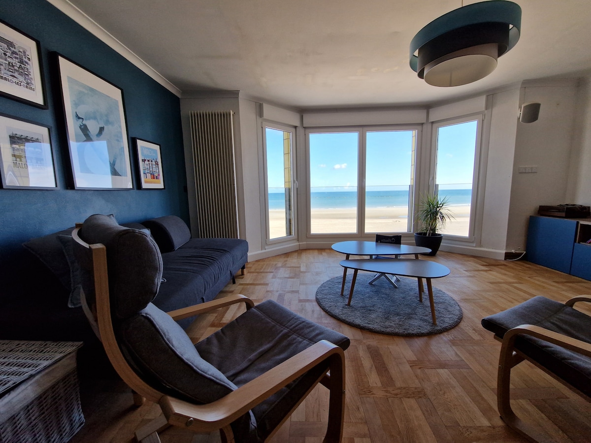 A spacious living area features large bay windows offering direct sea views. A modern sofa and two comfortable armchairs surround a circular coffee table, set on a soft area rug. Artwork adorns the walls, complementing the light and airy ambiance.