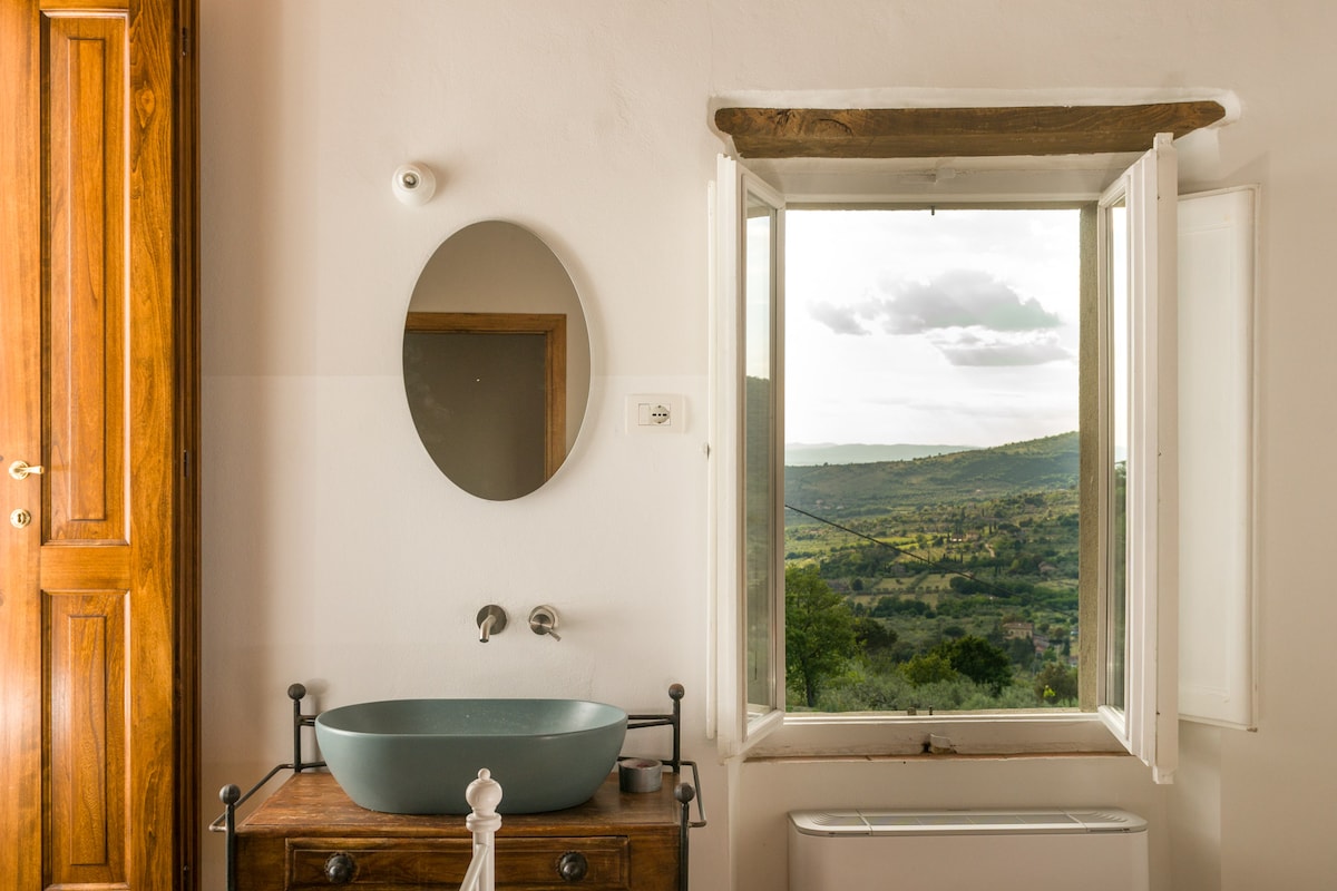 A bathroom features a round mirror above a modern basin on a wooden vanity. Natural light floods the space through a window that opens to reveal a scenic view of the countryside and rolling hills under a partly cloudy sky.