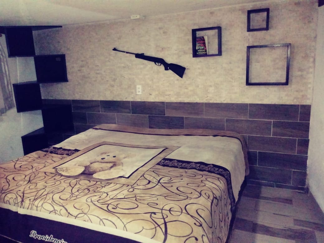 A cozy sleeping area features a king size bed adorned with a patterned blanket and a decorative bear motif. A wall-mounted rifle and empty frames add character to the neutral-toned walls, while shelving units provide storage space.