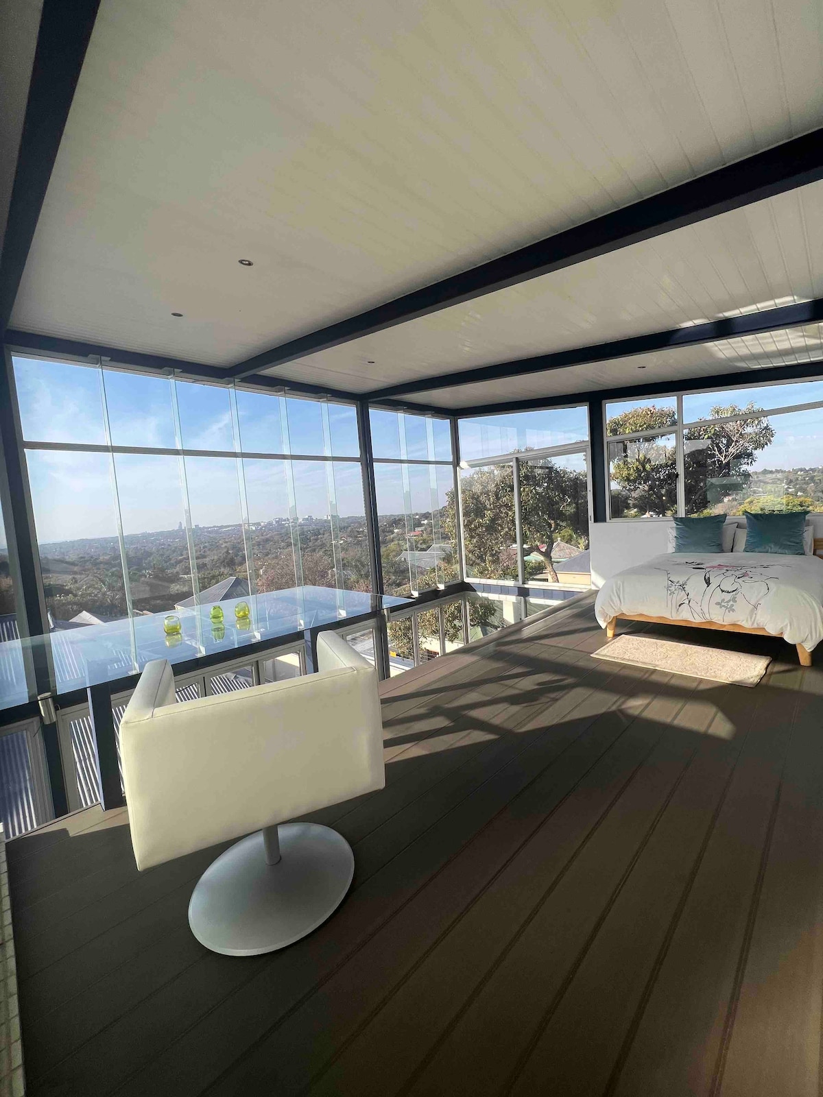 A bright bedroom features floor-to-ceiling windows showcasing expansive views of the cityscape. A spacious bed is adorned with soft linens, while a sleek white chair and table are positioned nearby, inviting relaxation in a light-filled environment.