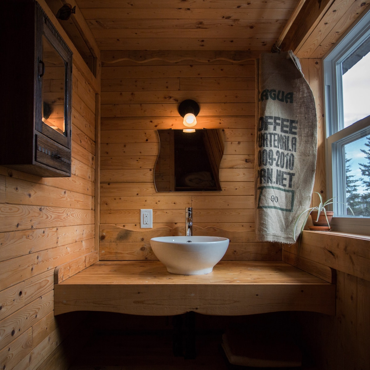 A wooden bathroom features warm tones from the natural wood paneling. A round basin sink sits on a handcrafted countertop, while a wall mirror reflects soft lighting from a vintage-style lamp. A burlap sack hanging by the window adds a rustic touch.