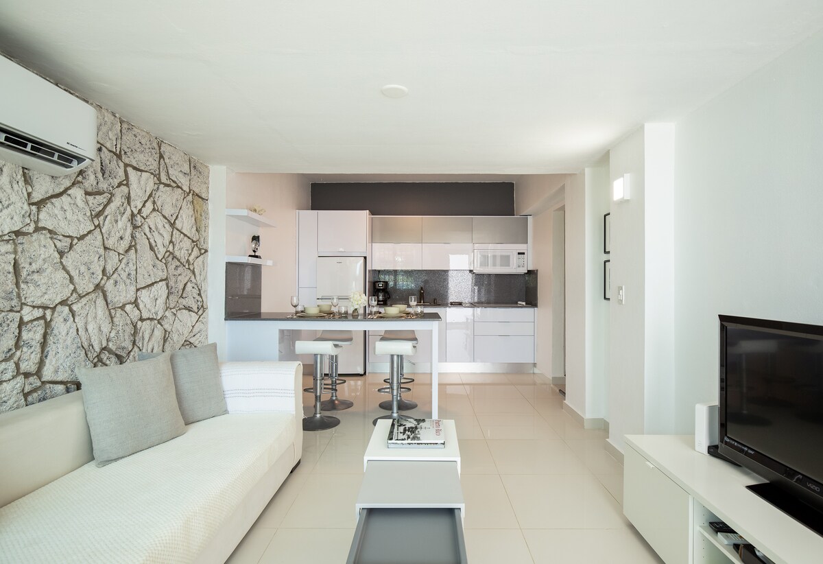The open-concept living area features a comfortable sofa and a flat-screen TV. A modern kitchen with sleek cabinetry and quartz countertops is visible in the background, alongside a dining space with two bar stools, creating a bright and inviting atmosphere.