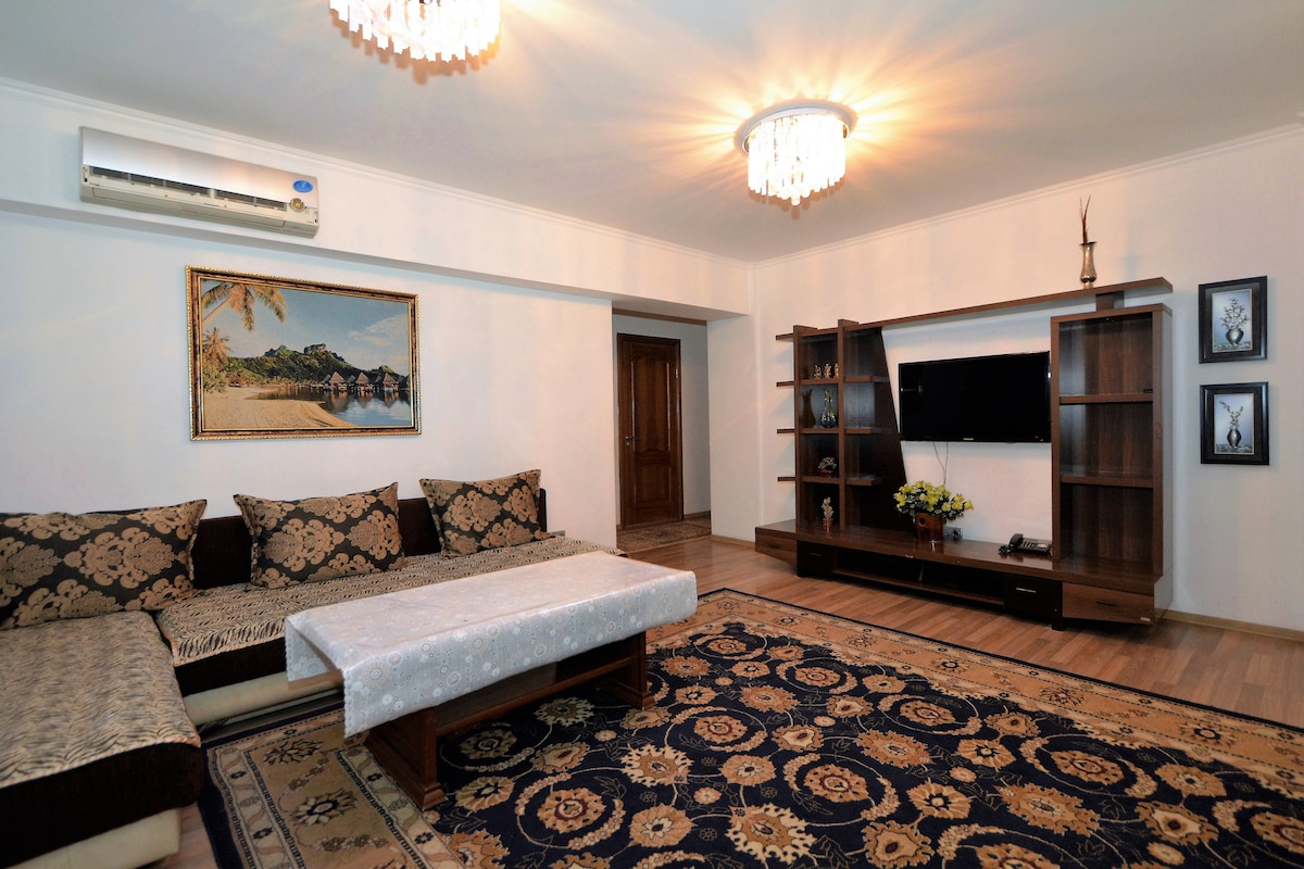 A spacious living area features elegant light fixtures and a detailed area rug. A comfortable seating arrangement is complemented by a decorative painting on the wall. The space includes a flat-screen television mounted on a wooden console, enhancing the overall comfort.