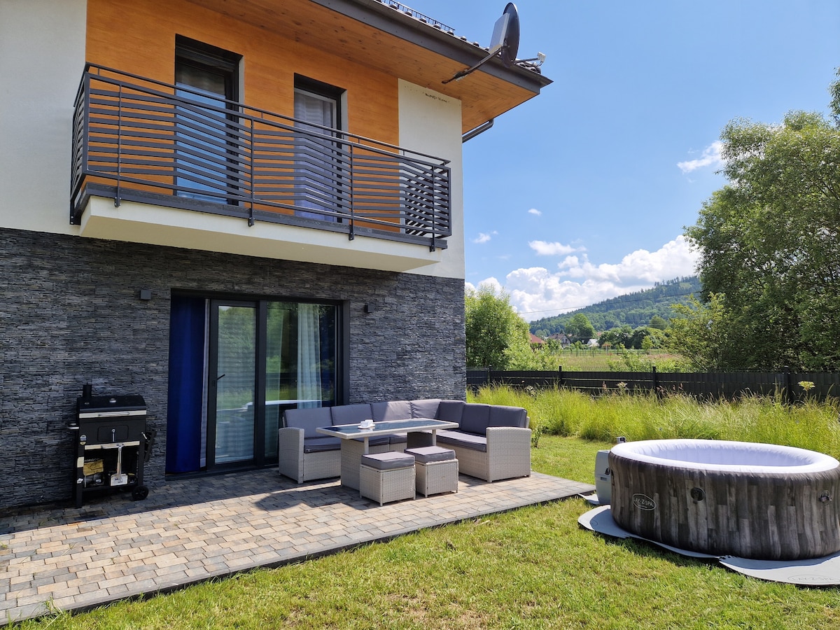 A private outdoor space features a comfortable seating area with a sectional sofa and a coffee table. A portable grill is positioned nearby. A hot tub is visible, surrounded by greenery, and the structure's upper balcony includes railings, complementing the scenic mountain backdrop.