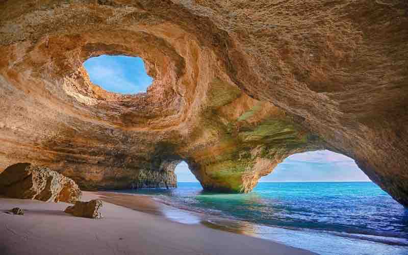 A scenic coastal cave features two circular openings in the rocky ceiling, allowing natural light to illuminate the sandy beach below. Gentle waves lap at the shore, while the turquoise water contrasts with the warm tones of the stone walls, adding to the serene environment.