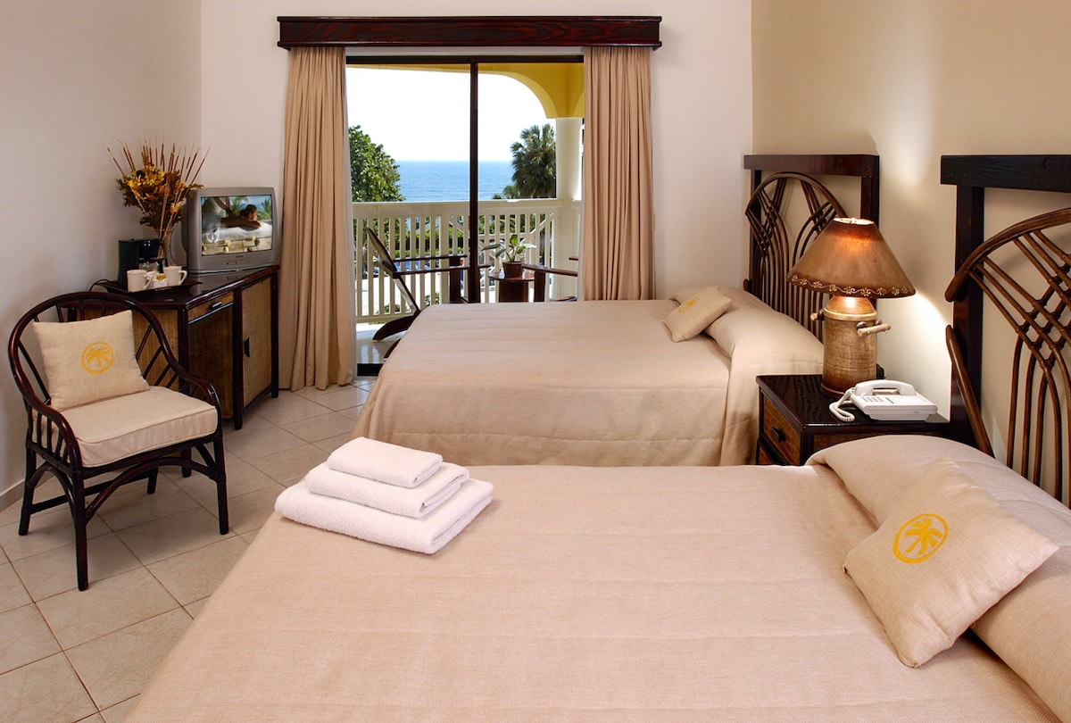 A well-appointed guest room features two spacious beds with neutral bedding. A balcony with ocean views is accessible through sliding glass doors. A seating area with a wicker chair and a small table complements a wooden dresser and lamp, creating a relaxing ambiance.