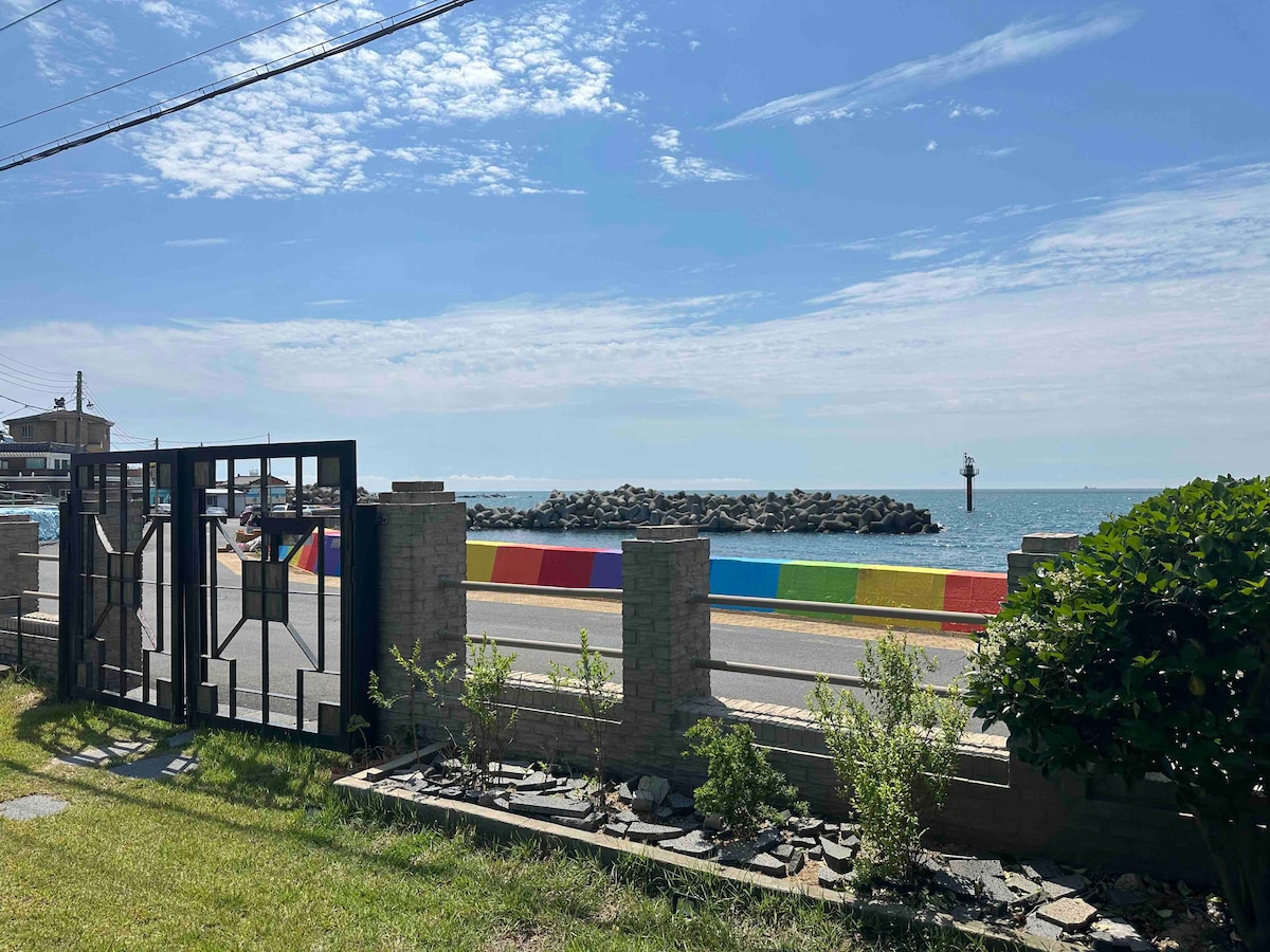 A view of the seaside is captured through a decorative gate, with vibrant rainbow-colored barriers lining the road. The calm water and the distant breakwater create a serene coastal atmosphere, complemented by a clear blue sky dotted with light clouds.
