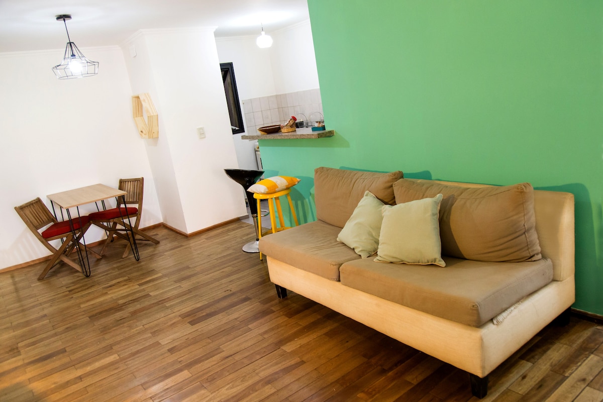 A living area features a brown couch with decorative cushions, accompanied by a small dining table with four folding chairs. The space is illuminated by overhead lighting, and a wood-paneled floor complements the fresh green accent wall.